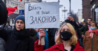 Person mit rotem Mantel hält ein Plakat mit der Aufschrift 'Vlast ne vyshe zakonov' während einer Demonstration.